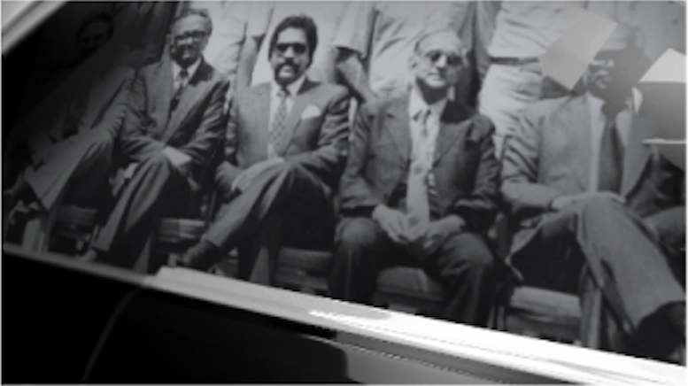 'Bad Boy Billionaires' title sequence style frame 6: close up of a black and white photograph, showing four Indian men in suits and sunglasses sat next to each other.