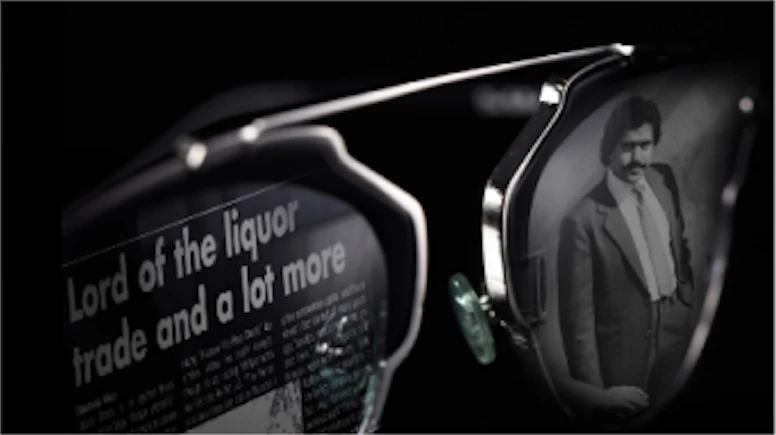 'Bad Boy Billionaires' title sequence style frame 8: close up of a pair of sunglasses, with a black and white image in each lense. The right lense shows a photo of a man in a suit. The left lense shows the headline of a newspaper article which reads 'Lord of the liquor trade and a lot more'