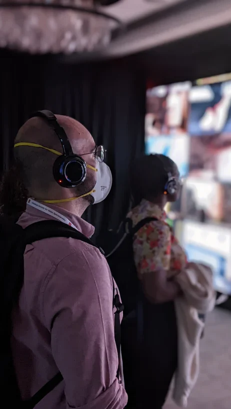 2 people wearing headphones watching our installation- 'tiles' of videos cover a whole wall which you can watch and listen to.