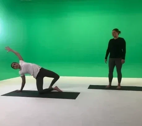 two instructors on exercise mats in front of a green screen. One is standing, the other is showing a yoga move.