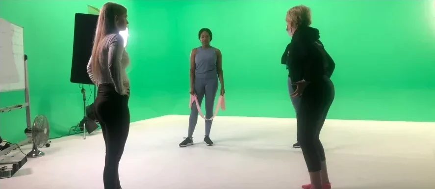 3 instructors standing in a circle, in front of the green screen. Some lighting equipment is in the background
