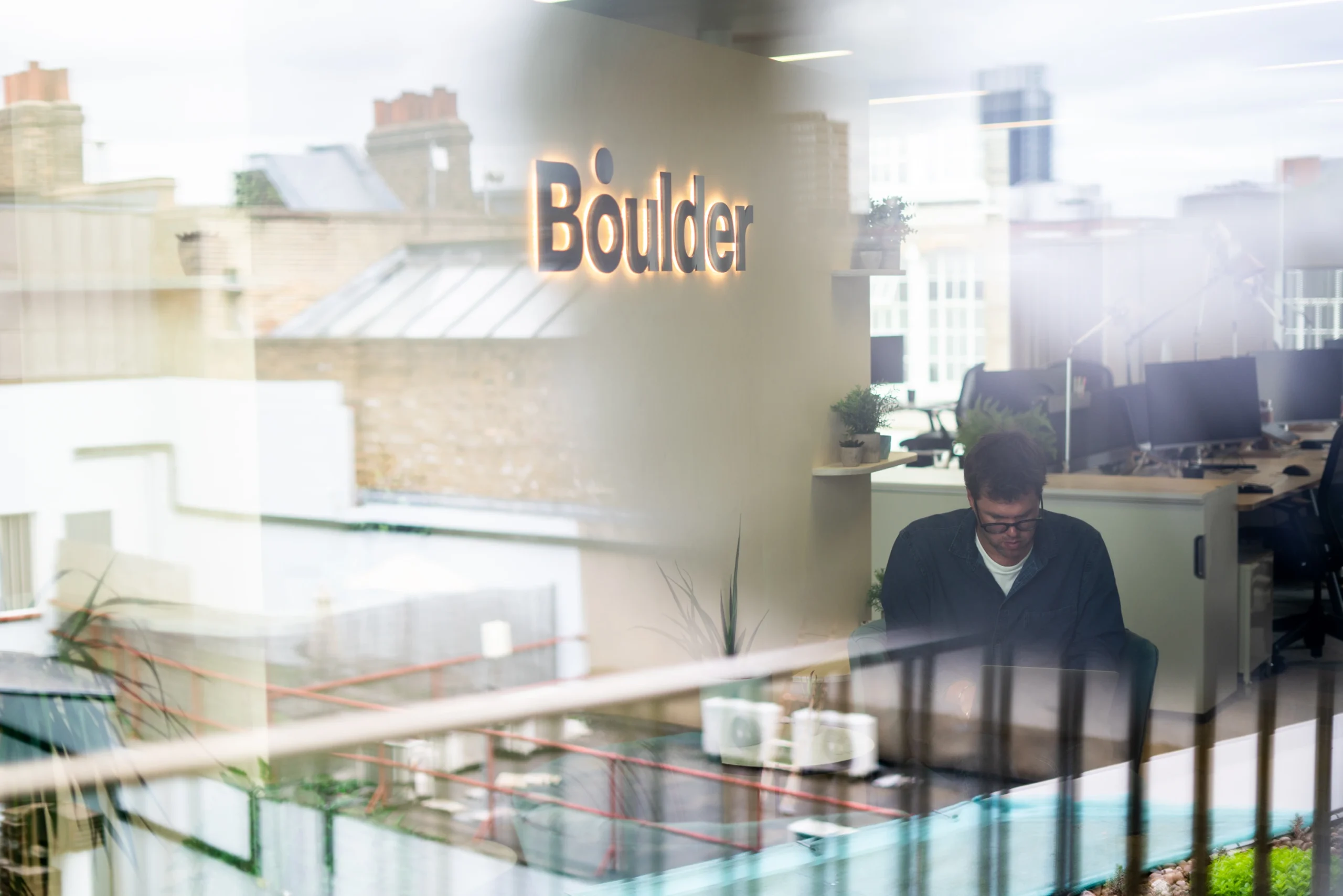 Photo taken through the office window, of the Managing Director sat in the office entryway underneath the 'Boulder' sign, working on his laptop.