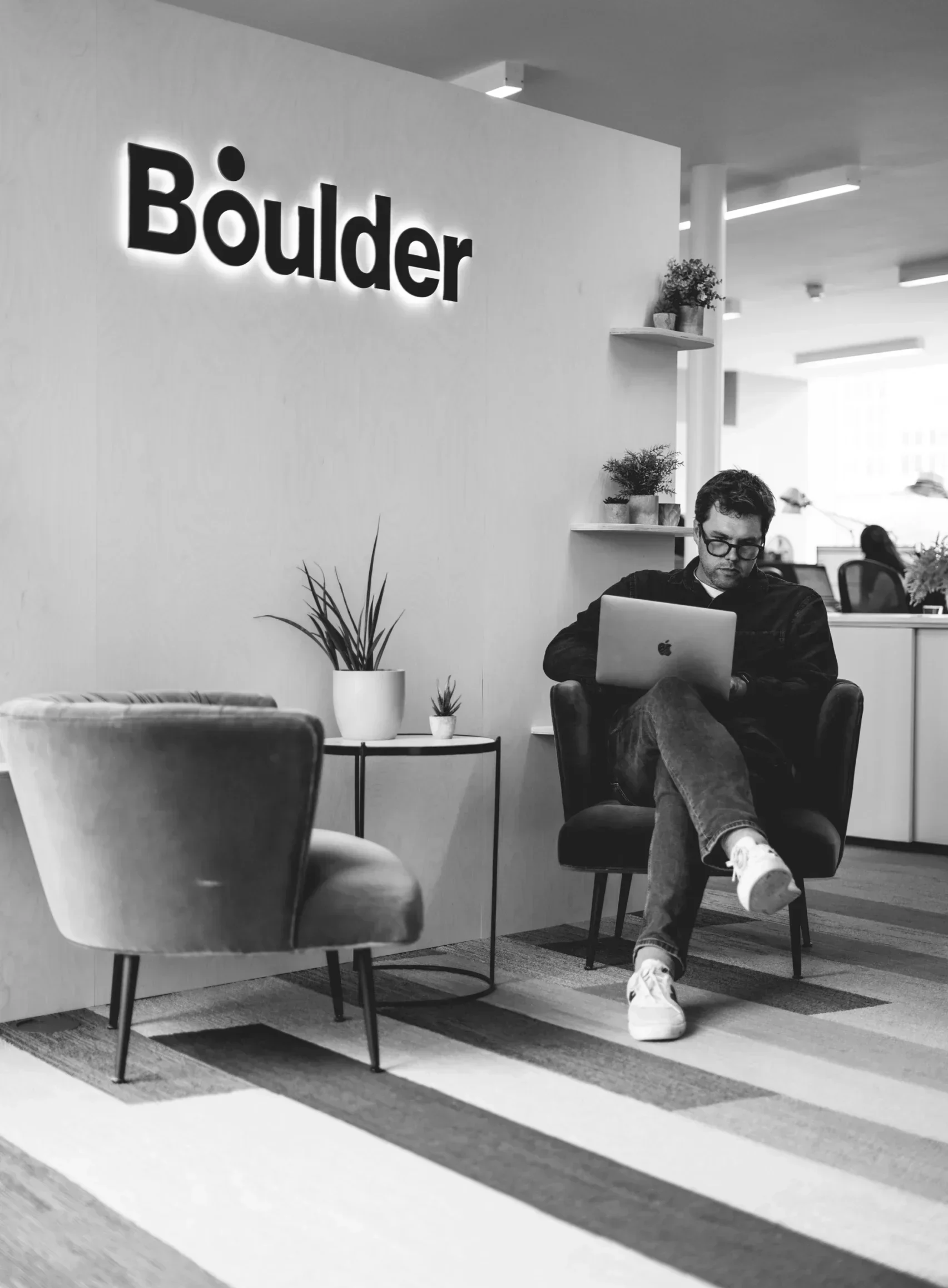 Black and white photo of our Managing Director working on his laptop, sat in the entrance of our office, with our 'Boulder' sign on the wall above him.